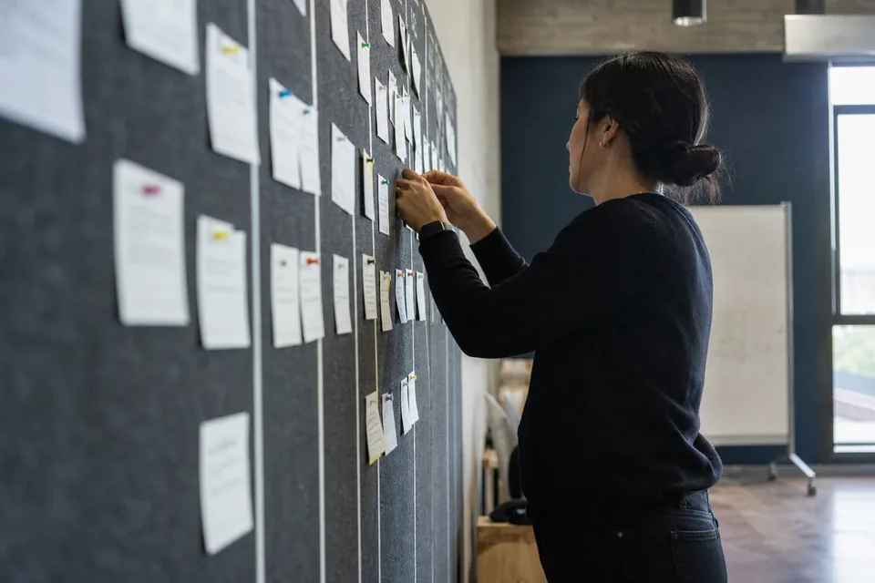 Marketer pinning printed quote cards onto a wall board sorted into columns, photographed from a low angle