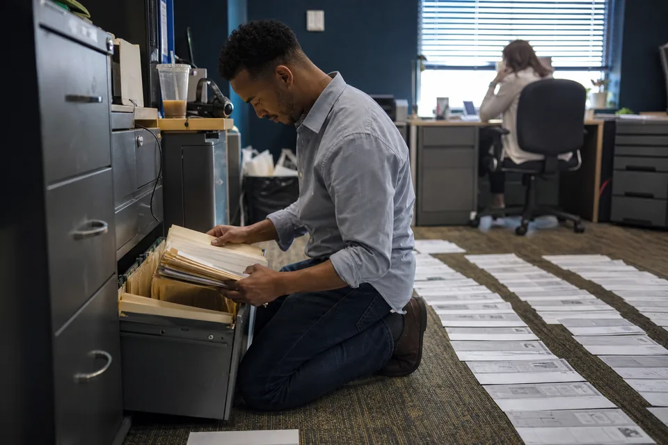 Customer success specialist kneeling at an open filing cabinet pulling folders of printed survey sheets, with rows of printouts laid out on the floor
