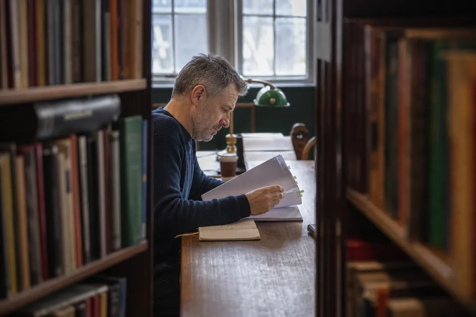 Content marketer reading a stack of printed guides at a library table and marking passages with tabs