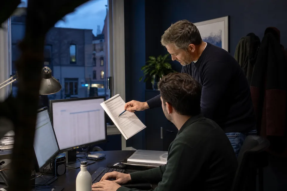 Agency owner and a colleague reviewing a printed client report at two monitors in a small agency office