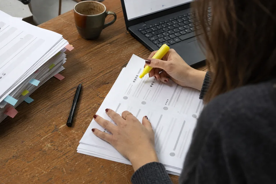Close-up of a content marketer's hands marking the strongest sentence of a printed customer review with a highlighter, a stack of review printouts sorted with coloured sticky tabs beside it