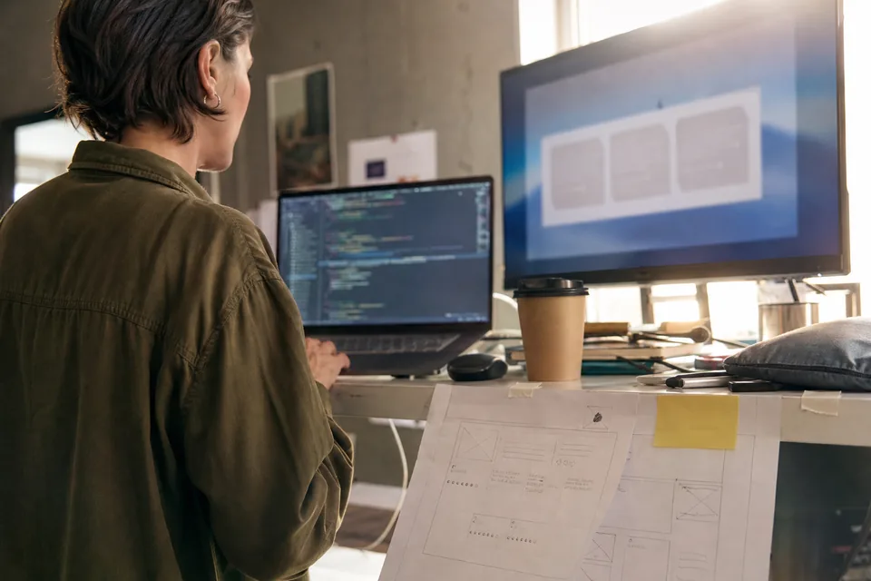 Front end developer pasting a small embed snippet into a page template on a laptop beside a printed wireframe